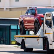 Broken red pickup car loaded onto white towing truck in public area for transportation to garage
